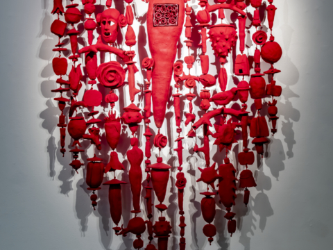 The Amulet Series 16 is a 48” x 57” low fired clay vertical wall hanging in red with figures of a human avatar, a face, thorny branches, geometric shapes, seed pods, birds, flames, flowers, and leaves.