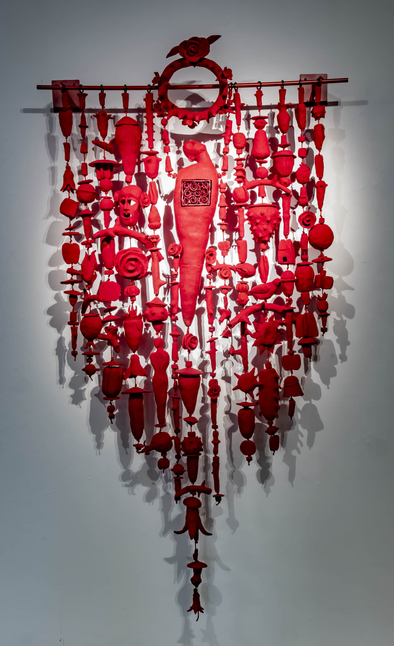 The Amulet Series 16 is a 48” x 57” low fired clay vertical wall hanging in red with figures of a human avatar, a face, thorny branches, geometric shapes, seed pods, birds, flames, flowers, and leaves.