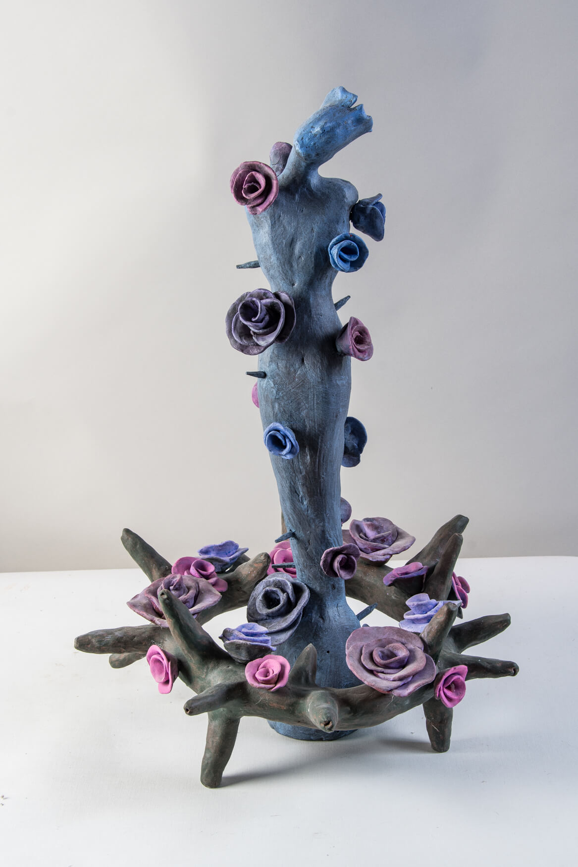 Twilight is mixed media on low fired clay. It has a brown crown of thorns covered with blue, purple, and pink flowers with a blue human figure rising vertically from the center that is also covered in flowers and thorns.