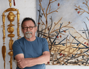 Photo of artist Paul Medina. He is wearing a blue shirt and glasses. On his left is a gold amulet hanging and on the right is a briar series collage.