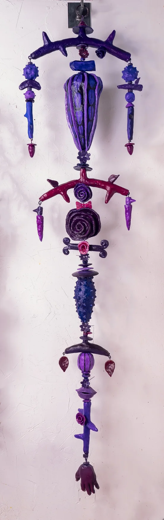 Hanging mixed media sculpture titled 'Aubergine' by Paul Medina, featuring a vertical arrangement of abstract and organic forms in purple and deep red hues. The sculpture includes various shapes resembling branches, a spiral, and a hand at the bottom, all interconnected and suspended against a light background.