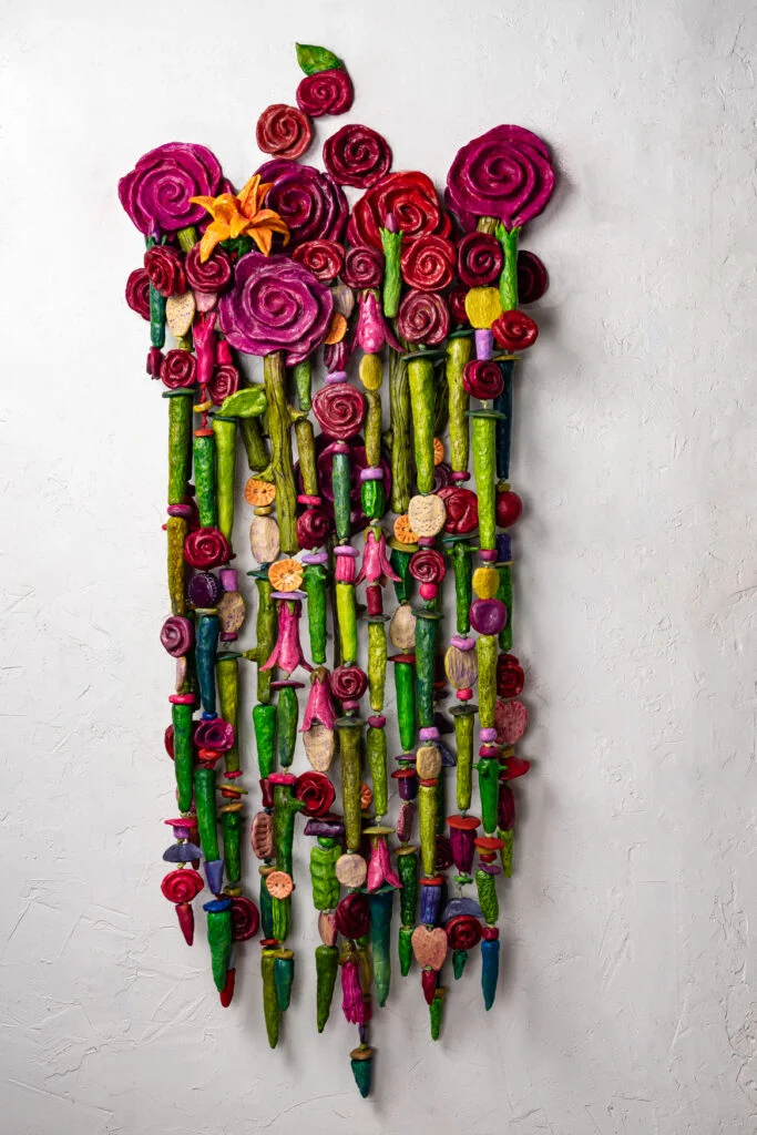 Bouquet For Grace by Oklahoma born mixed media artist is a colorful vertical wall sculpture featuring layered, abstract flowers in vibrant shades of red, pink, and green, with a single orange lily at the top.