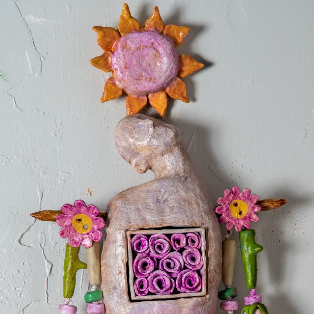 Love Never Dies is a 48" x 12" x 7" hanging wall amulet of mixed media on fired clay, steel, copper, and nylon cord. the image is a detail of a human figure balancing a sunflower on its head with a box of roses over its heart, The colors are pastel tones in pinks, purples, orange and greens.